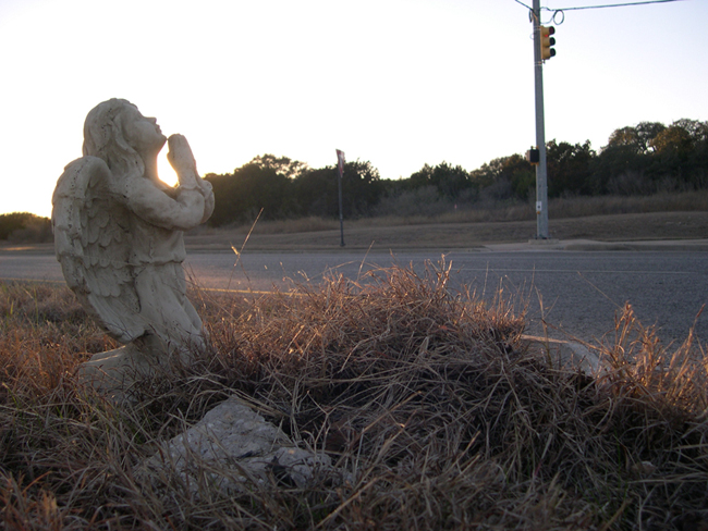 The setting sun glorifies the angel in a side view of the scene.  A piece of broken curb is nestled in the grass.