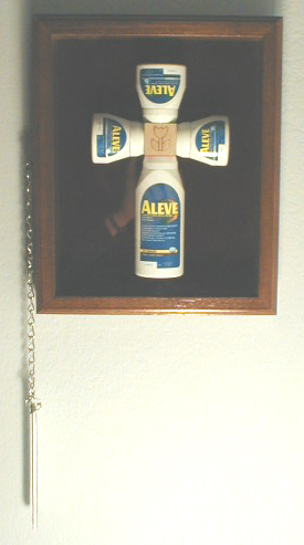 The cross is formed from 4 OTC pain-pill bottles in 3 different sizes, secured by screws through lids into central wooden play-block, with pyrographed flower icon prominent.  Cross is secured in glass-fronted wooden display case lined with burgandy velvet and equipped with external chain and breaker bar, like a fire extinguisher cabinet.