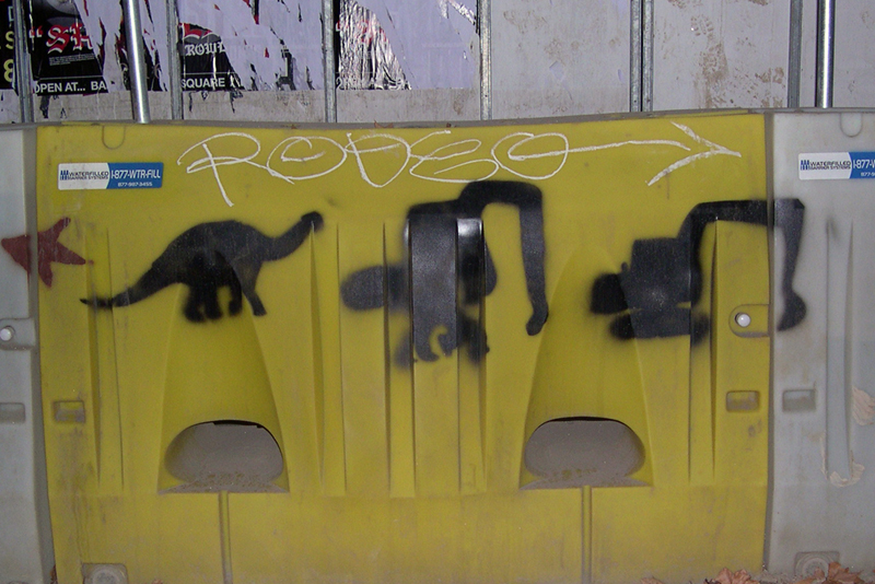 3 spraypainted black glyphs on a yellow barrier morph from a brontosaur to an excavator.