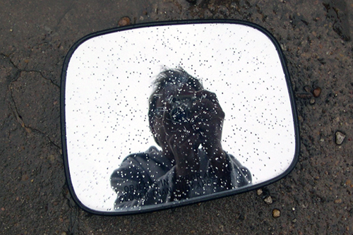 The broken-off but intact side-view mirror of an automobile lies face-up in the street, the author's face and camera visible in the reflection off its surface.