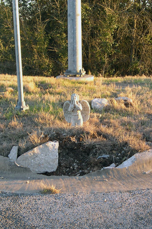 A concrete angel raises its hands to heaven above a section of demolished curb at a roadside memorial in Austin, TX.